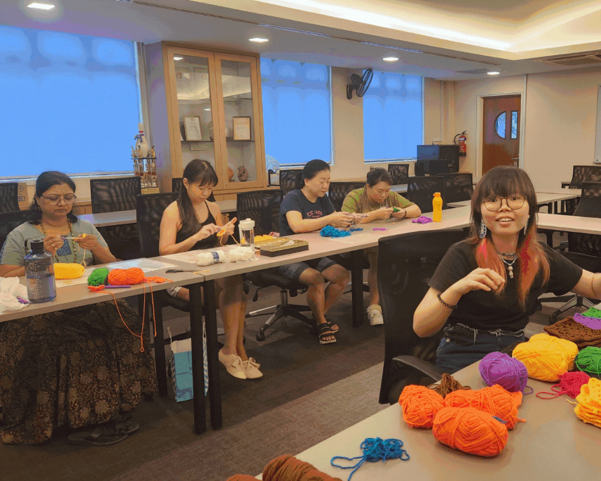 Five women sit around a table crocheting, with balls of brightly coloured yarn in the foreground.